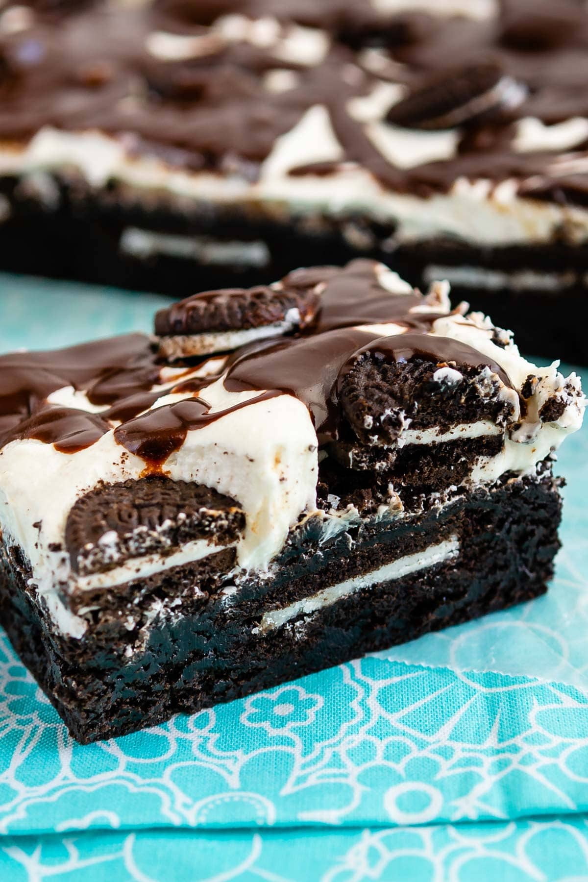 square slice of brownies with white frosting and chocolate drizzle on top sitting on a teal cloth