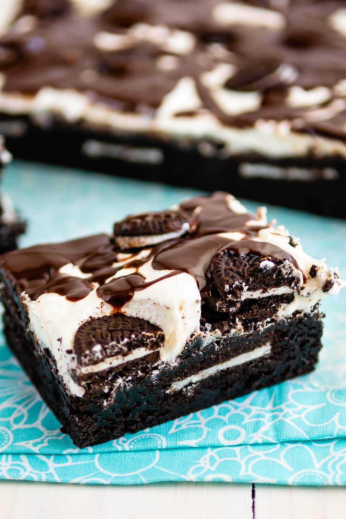 square slice of brownies with white frosting and chocolate drizzle on top sitting on a teal cloth