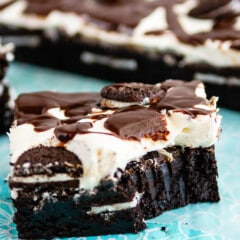 square slice of brownies with white frosting and chocolate drizzle on top sitting on a teal cloth