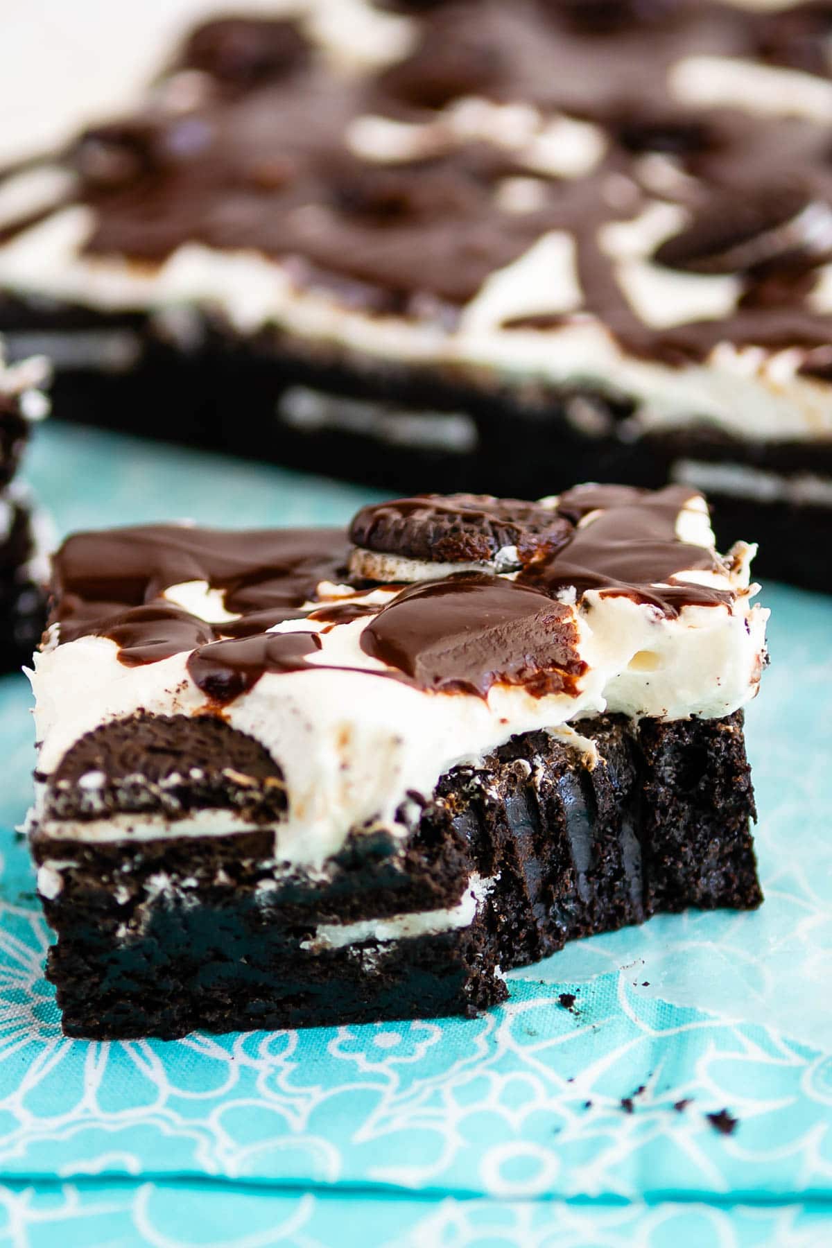 square slice of brownies with white frosting and chocolate drizzle on top sitting on a teal cloth