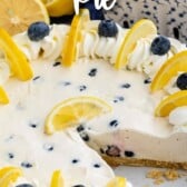 Lemon blueberry pie with one slice cut out of dish and blueberries and lemons in background and recipe title on top of photo