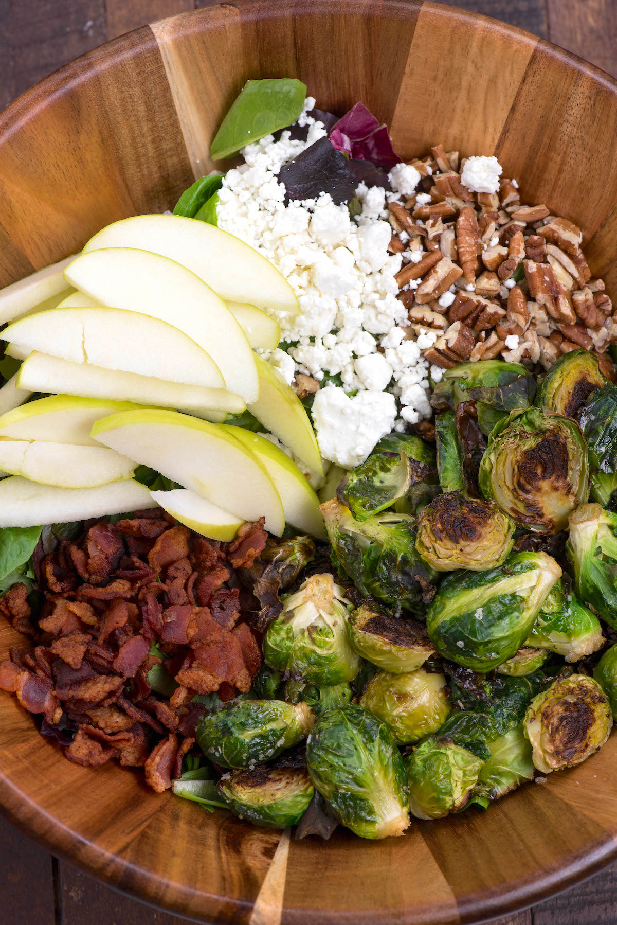 brussel sprout salad with all the ingredients including apples, Brussel sprouts, cheese, and bacon separated in a wooden bowl