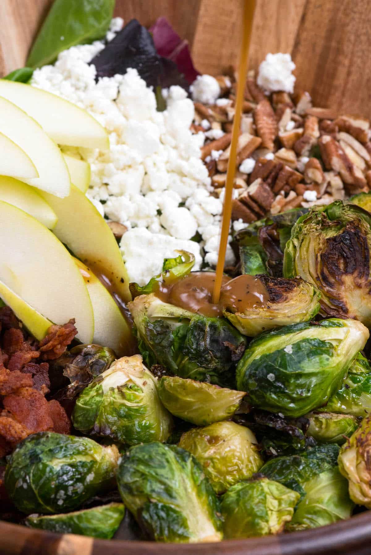 brussel sprout salad with all the ingredients including apples, Brussel sprouts, cheese, and bacon separated in a wooden bowl with dressing poured on top