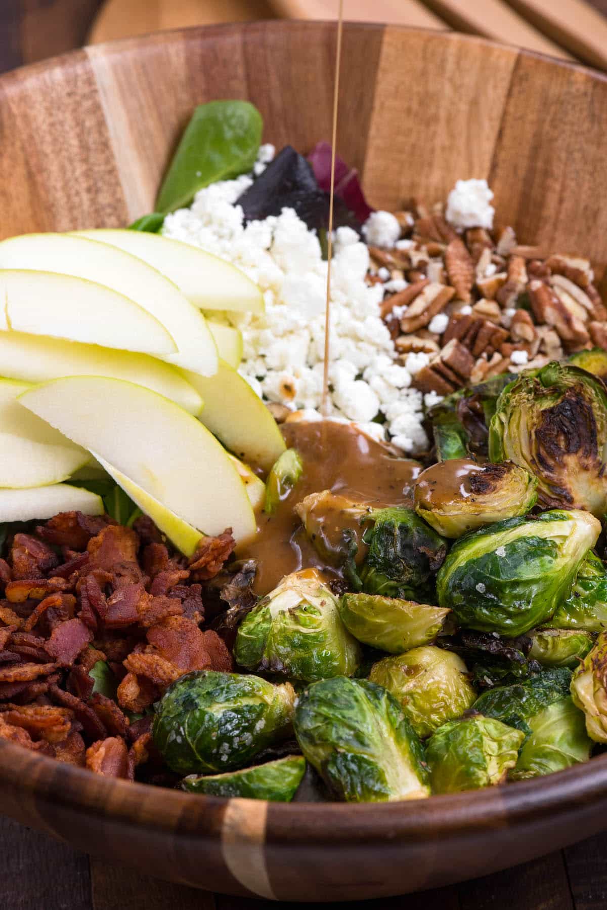 brussel sprout salad with all the ingredients including apples, Brussel sprouts, cheese, and bacon separated in a wooden bowl with dressing poured on top