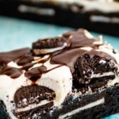 square slice of brownies with white frosting and chocolate drizzle on top sitting on a teal cloth with words on the photo