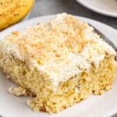A close-up of a slice of banana cake—one of the best banana recipes—with light, fluffy frosting and crumbs on top, served on a white plate with a fork. Bananas and another plate appear in the background.