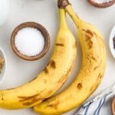 Two ripe bananas rest on a white surface, surrounded by bowls of chocolate chips, brown sugar, salt, cinnamon, and a striped cloth—perfect ingredients for delicious banana recipes.
