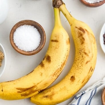 Two ripe bananas rest on a white surface, surrounded by bowls of chocolate chips, brown sugar, salt, cinnamon, and a striped cloth&mdash;perfect ingredients for delicious banana recipes.