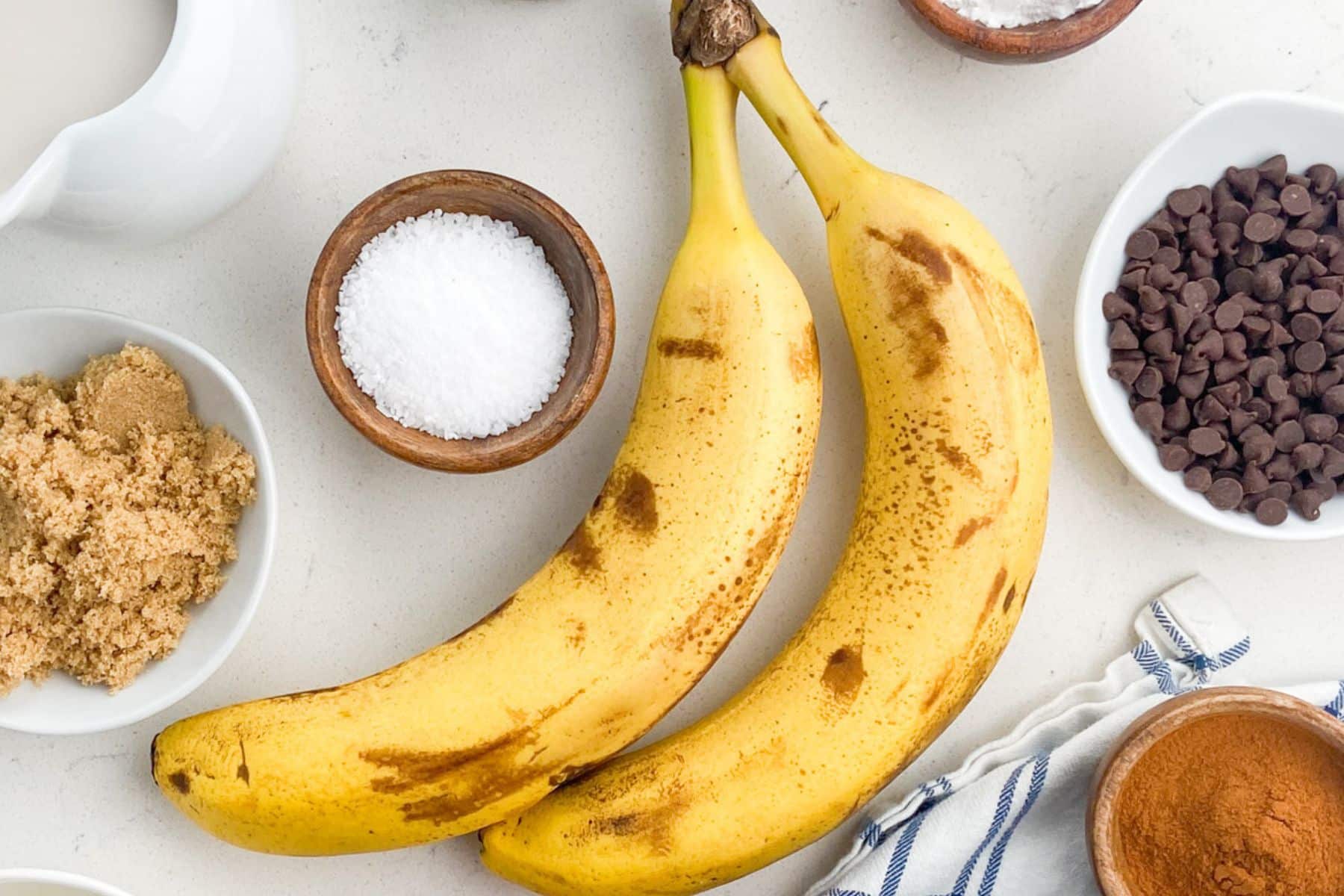 Two ripe bananas rest on a white surface, surrounded by bowls of chocolate chips, brown sugar, salt, cinnamon, and a striped cloth—perfect ingredients for delicious banana recipes.