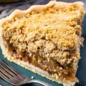 A close-up of a slice of Dutch apple pie on a dark green plate, with a fork beside it. The pie has a crumbly golden topping and flaky crust, revealing the tender apple filling inside.