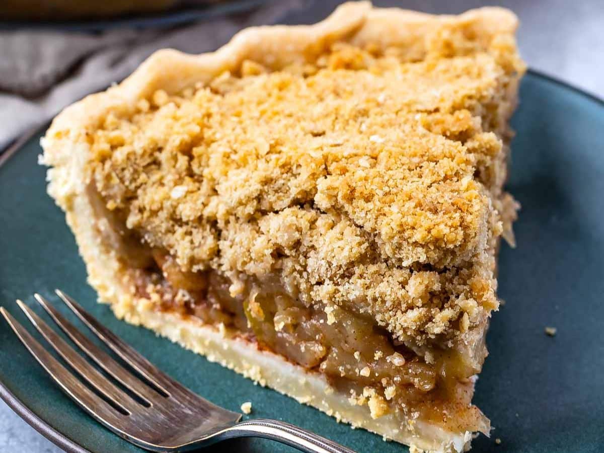 Gros plan d'une tranche de tarte aux pommes hollandaise sur une assiette vert foncé, avec une fourchette à côté. La tarte a une garniture dorée et friable et une croûte feuilletée, révélant la tendre garniture aux pommes à l'intérieur.