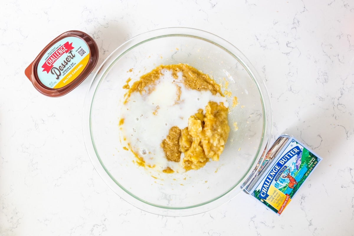 banana bread batter with milk in glass bowl on counter with butter and dessert spread.