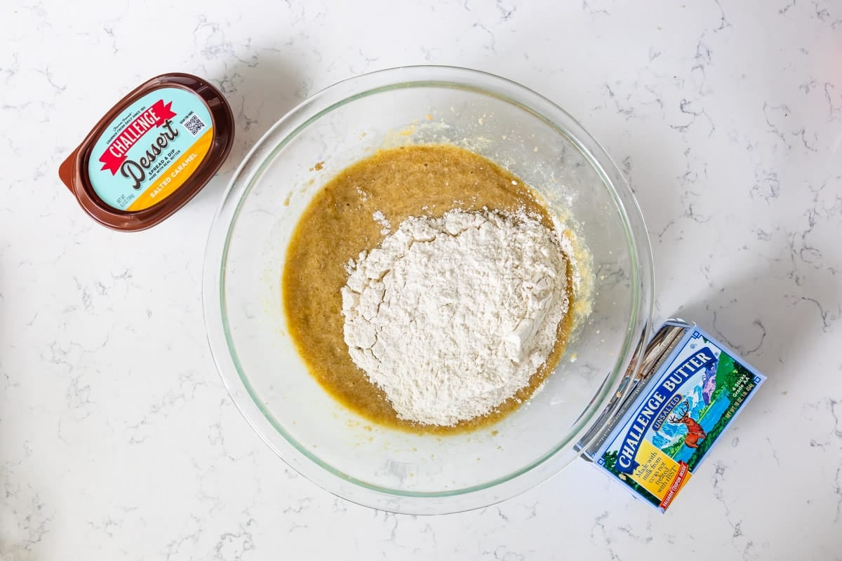 banana bread batter with flour in glass bowl on counter with butter and dessert spread.