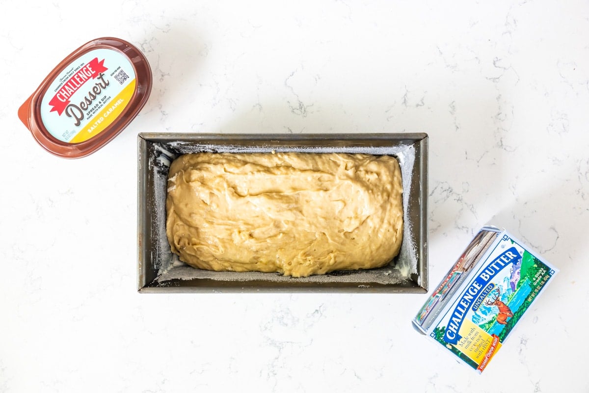 banana bread batter in loaf pan on counter with butter and dessert spread.