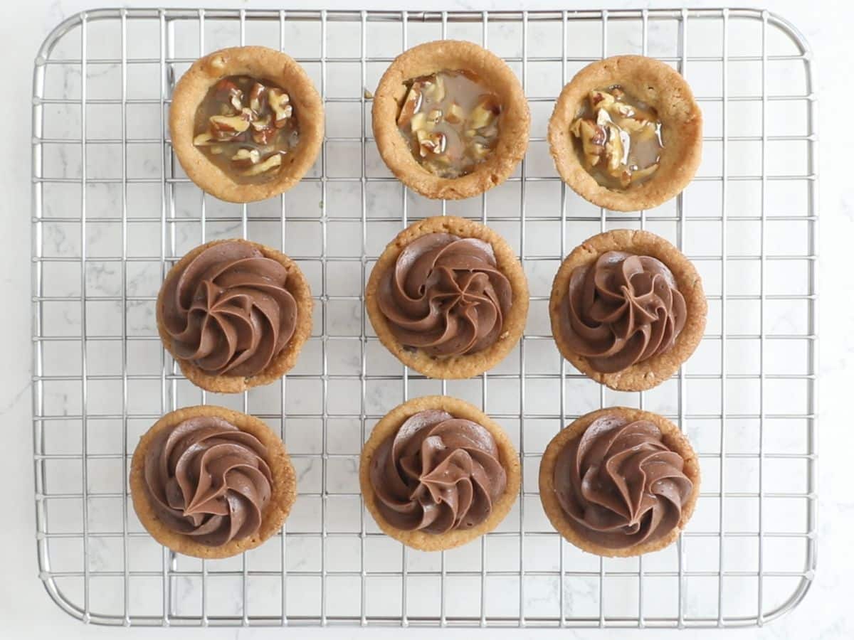 cookie cups on rack with caramel and pecans inside and 6 of them frosted with chocolate frosting.