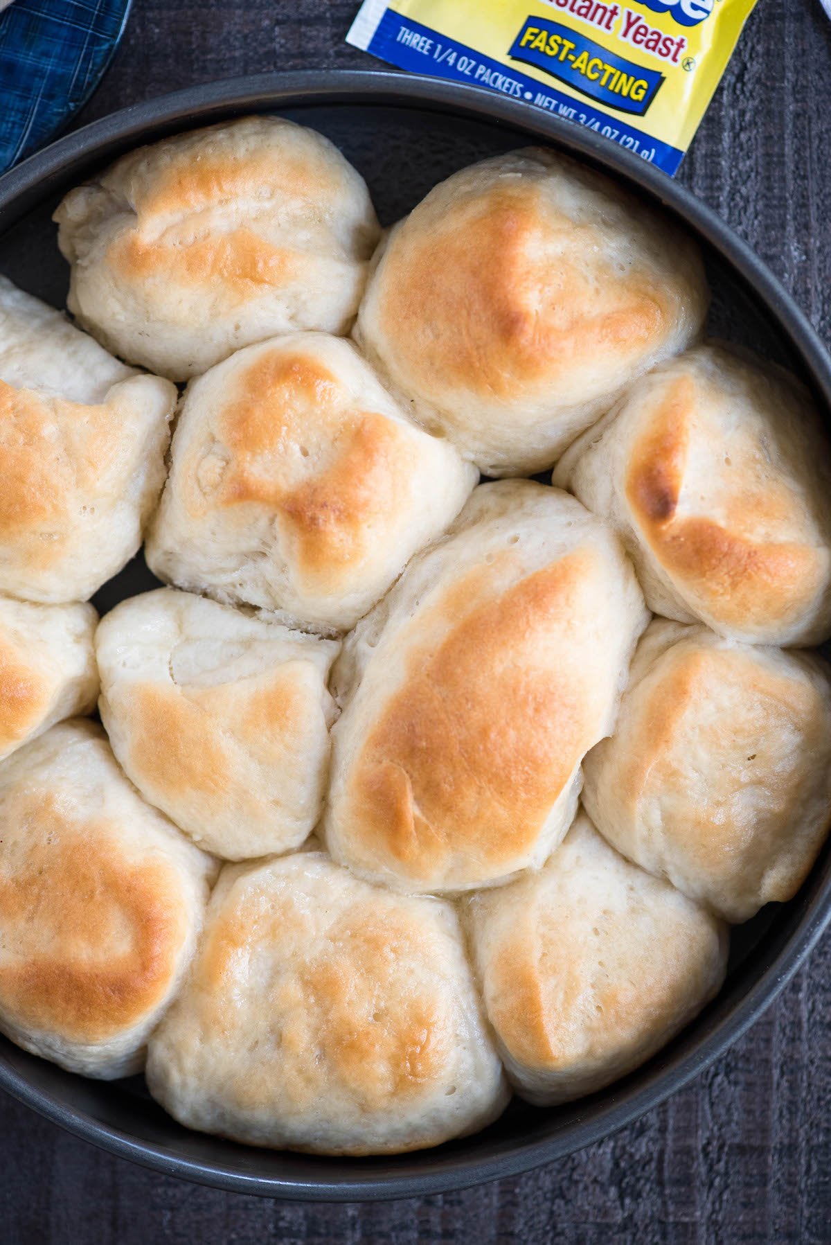 A round baking pan filled with freshly baked, golden-brown dinner rolls showcases the results of a cherished dinner roll recipe. The rolls, slightly irregular in shape, are closely packed together, with a packet of active dry yeast partially visible in the top right corner of the image.