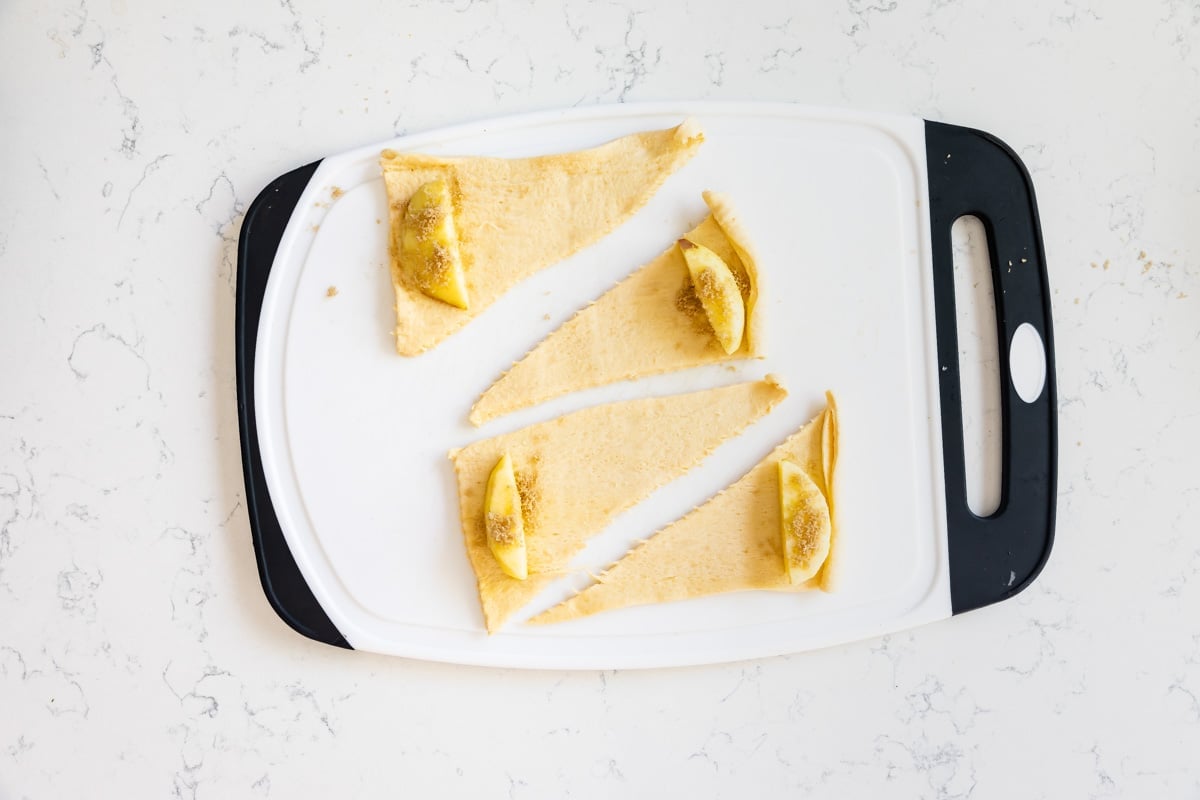 4 crescent rolls unrolled on white cutting board with apple slice an brown sugar.