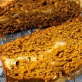 Two thick slices of moist pumpkin cream cheese bread with visible swirls of cream cheese rest on a metal cooling rack, highlighting the bread’s golden brown color and soft texture.