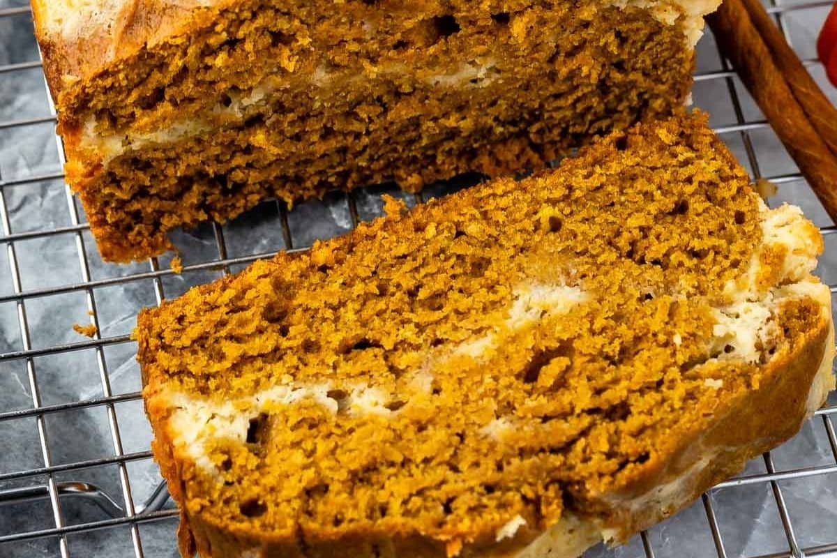 Two thick slices of moist pumpkin cream cheese bread with visible swirls of cream cheese rest on a metal cooling rack, highlighting the bread’s golden brown color and soft texture.