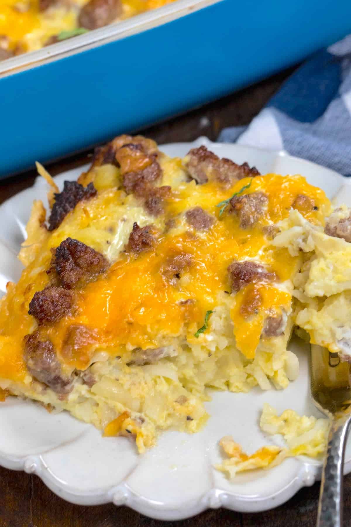 A serving of cheesy hashbrown breakfast casserole with sausage sits on a white plate beside a fork, with a blue baking dish and a striped napkin in the background.