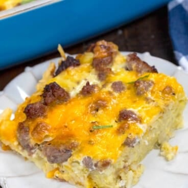 A slice of cheesy hashbrown breakfast casserole with sausage and eggs sits on a white plate, with more casserole visible in a blue baking dish in the background.