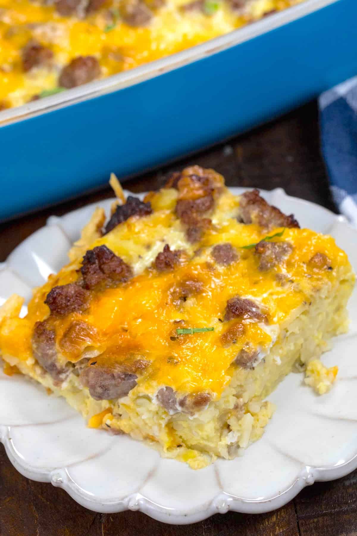 A slice of cheesy hashbrown breakfast casserole with sausage and eggs sits on a white plate, with more casserole visible in a blue baking dish in the background.
