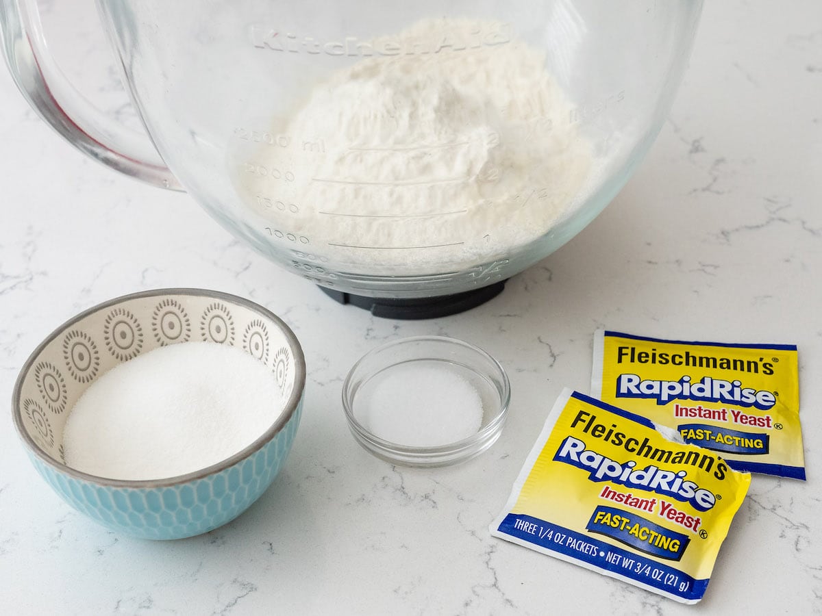 A mixing bowl with flour, a small glass dish, a patterned bowl of sugar, and two packets of Fleischmann’s RapidRise instant yeast await on a white countertop—perfect for making pumpkin spice cinnamon rolls.