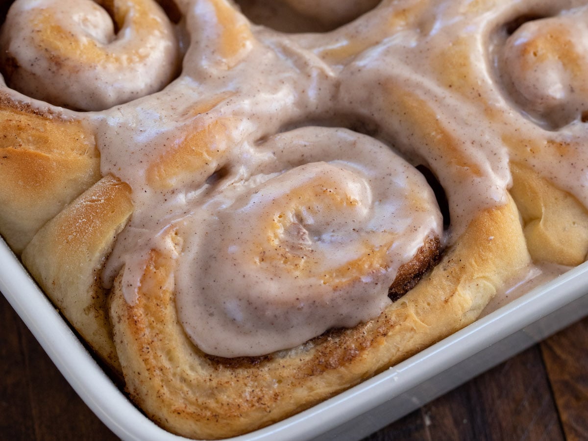 Close-up of freshly baked pumpkin spice cinnamon rolls in a white baking dish, topped with creamy, drizzled icing. The rolls are golden-brown and swirled, with visible cinnamon and pumpkin spice filling.