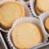 Round, golden butter cookies are arranged in white paper liners on a baking tray. These cookies, made from a classic butter cookies recipe, have a slightly crumbly texture and light, golden-brown edges.