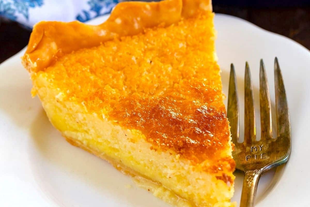 A slice of golden brown buttermilk pie with a flaky crust rests on a white plate, next to a fork engraved with the words MY PIE.