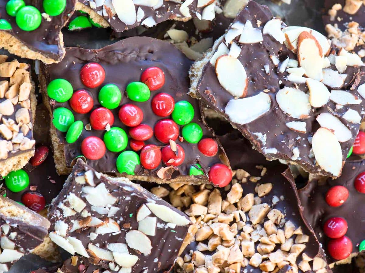 Assorted pieces of chocolate toffee bark and graham cracker toffee, topped with red and green candy-coated chocolates, sliced almonds, and crushed toffee pieces. The treats are arranged in a festive, colorful pile.