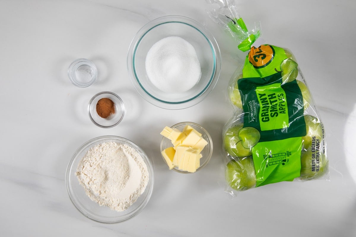 Top-down view of baking ingredients for Apple Slab Pie on a white surface, featuring a bag of Granny Smith apples, cubed butter, flour, sugar, cinnamon, and salt.
