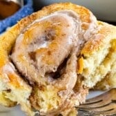 A close-up of a pumpkin spice cinnamon roll topped with melting cinnamon-sugar icing, showing its soft, fluffy texture. A fork rests beside the pastry on a white plate.