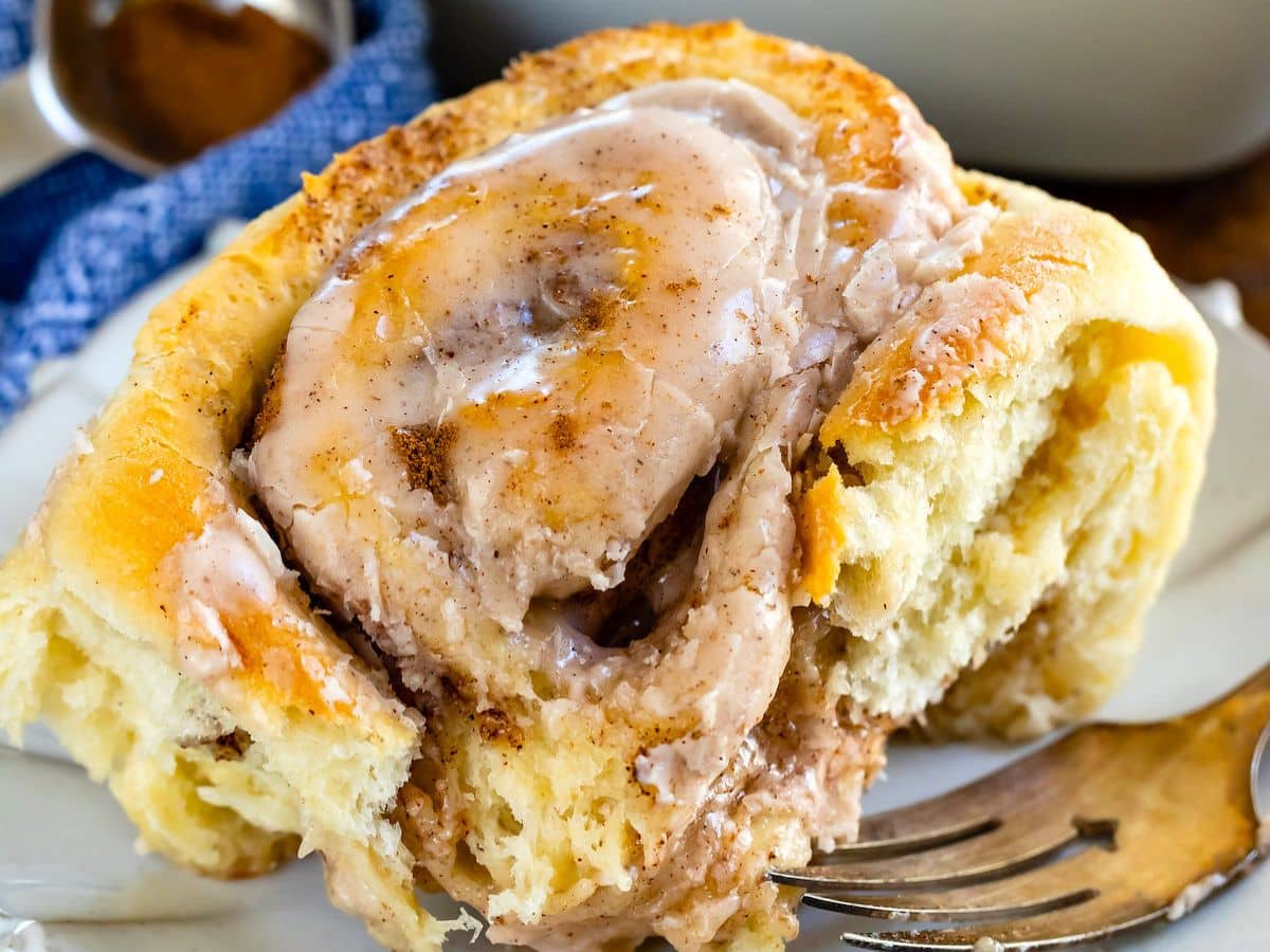 A close-up of a pumpkin spice cinnamon roll topped with melting cinnamon-sugar icing, showing its soft, fluffy texture. A fork rests beside the pastry on a white plate.