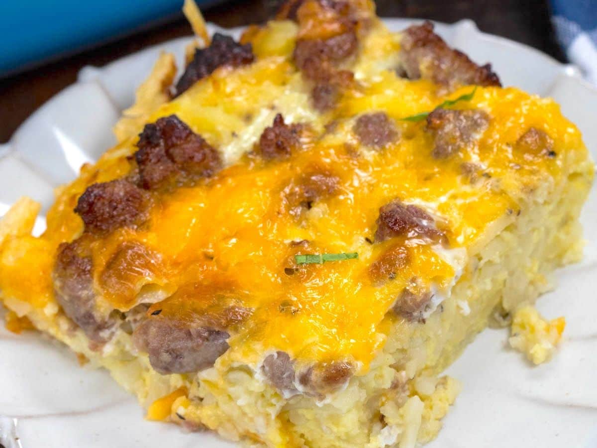 A slice of hashbrown breakfast casserole on a white plate, topped with melted cheese and pieces of sausage.