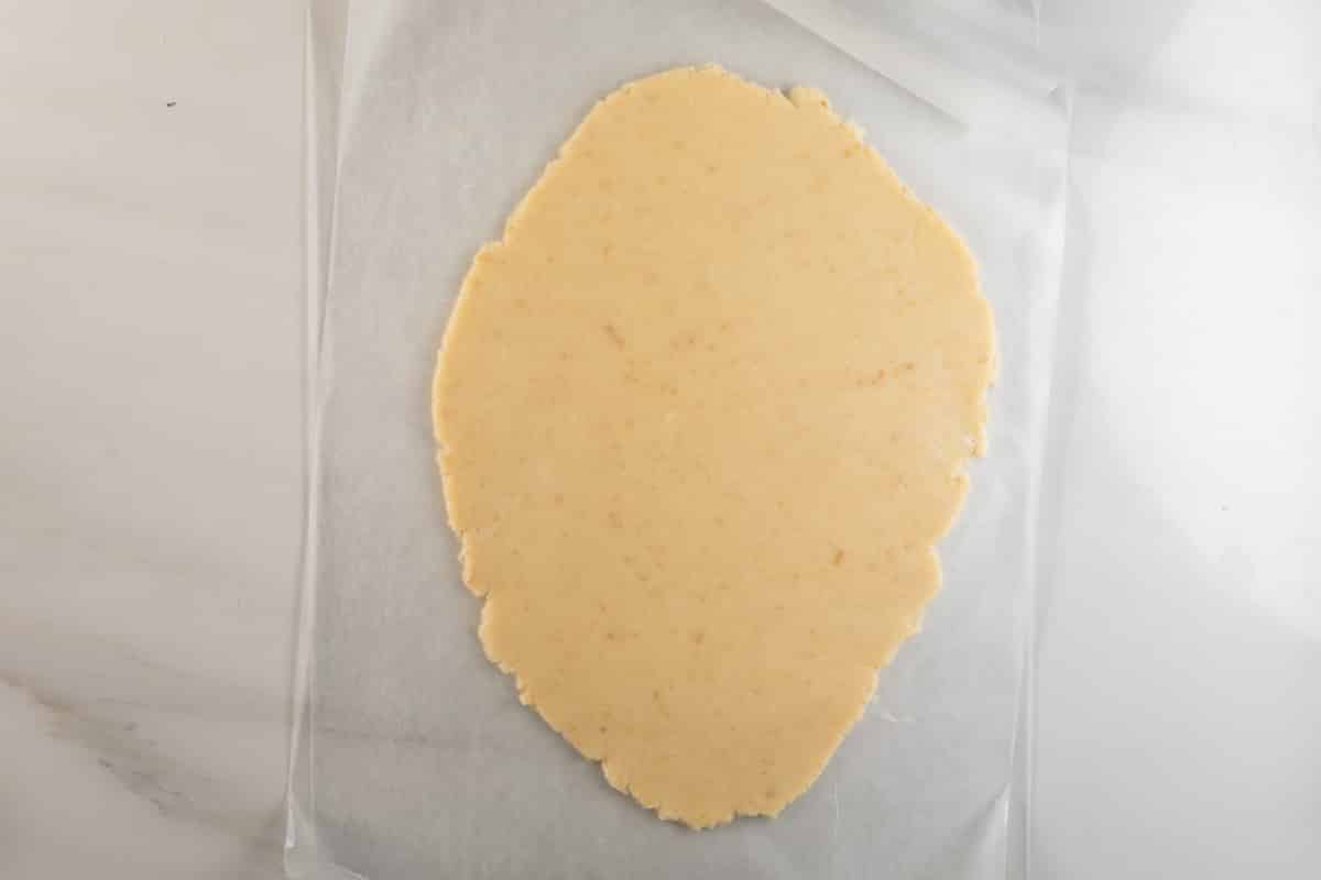 A sheet of pale, rolled-out dough sits on parchment paper atop a white surface, ready to be cut or shaped for baking the perfect shortbread cookies recipe.