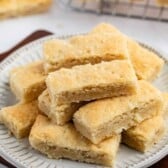 A plate of rectangular golden shortbread cookies, made from a classic shortbread cookies recipe, stacked on top of each other, with more cookies cooling on a wire rack in the background.