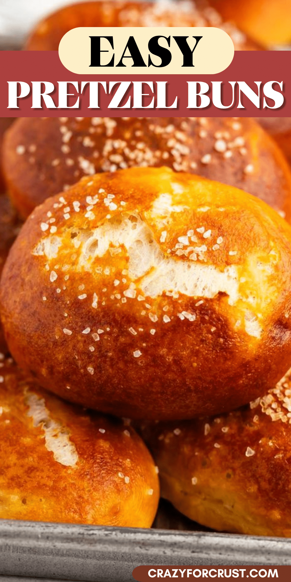 A close-up of golden-brown Easy Pretzel Rolls topped with coarse salt, stacked together on a tray. Text on image reads Easy Pretzel Buns and crazyforcrust.com.