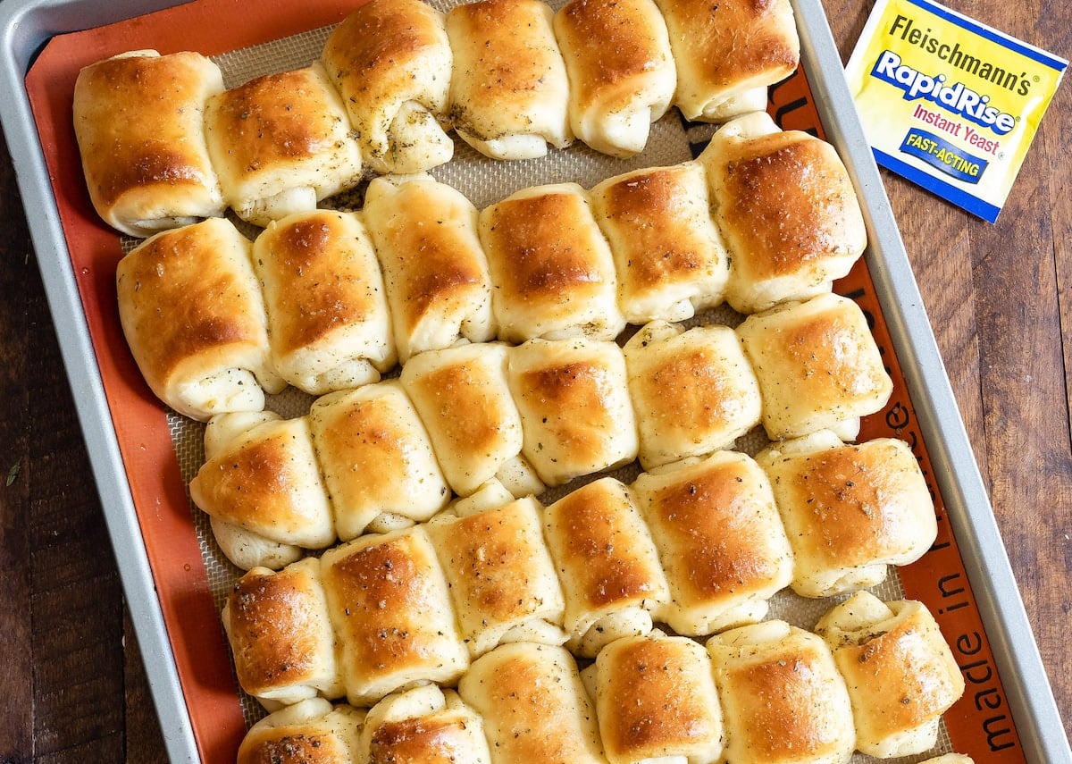 A baking sheet filled with golden-brown, freshly baked garlic rolls sits next to a packet of Fleischmann’s RapidRise Instant Yeast.