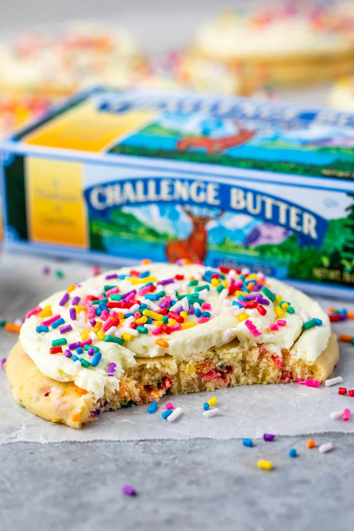 cookies with frosting and rainbow sprinkles on parchment with bite missing and butter box behind