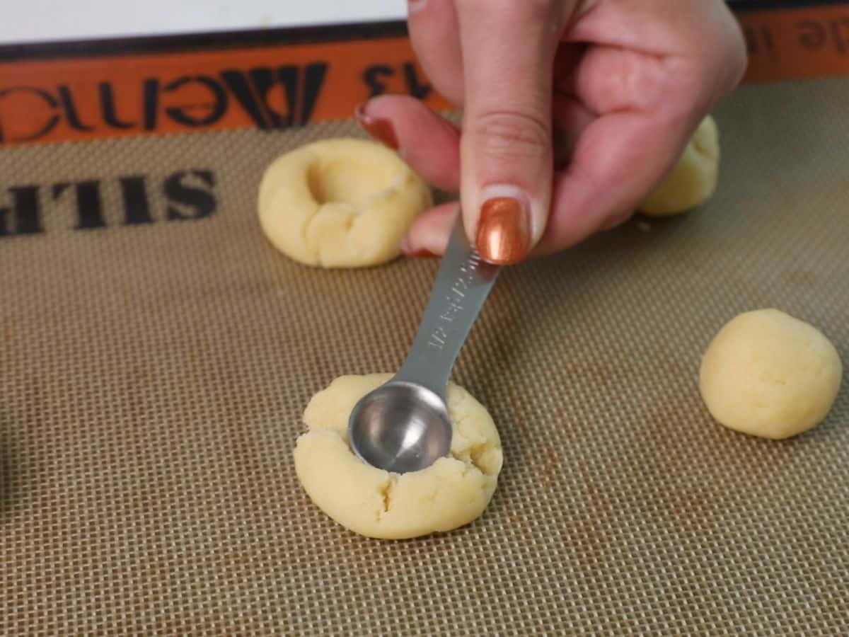 cookie dough balls on silat with hand pressing a measuring spoon into the ball.
