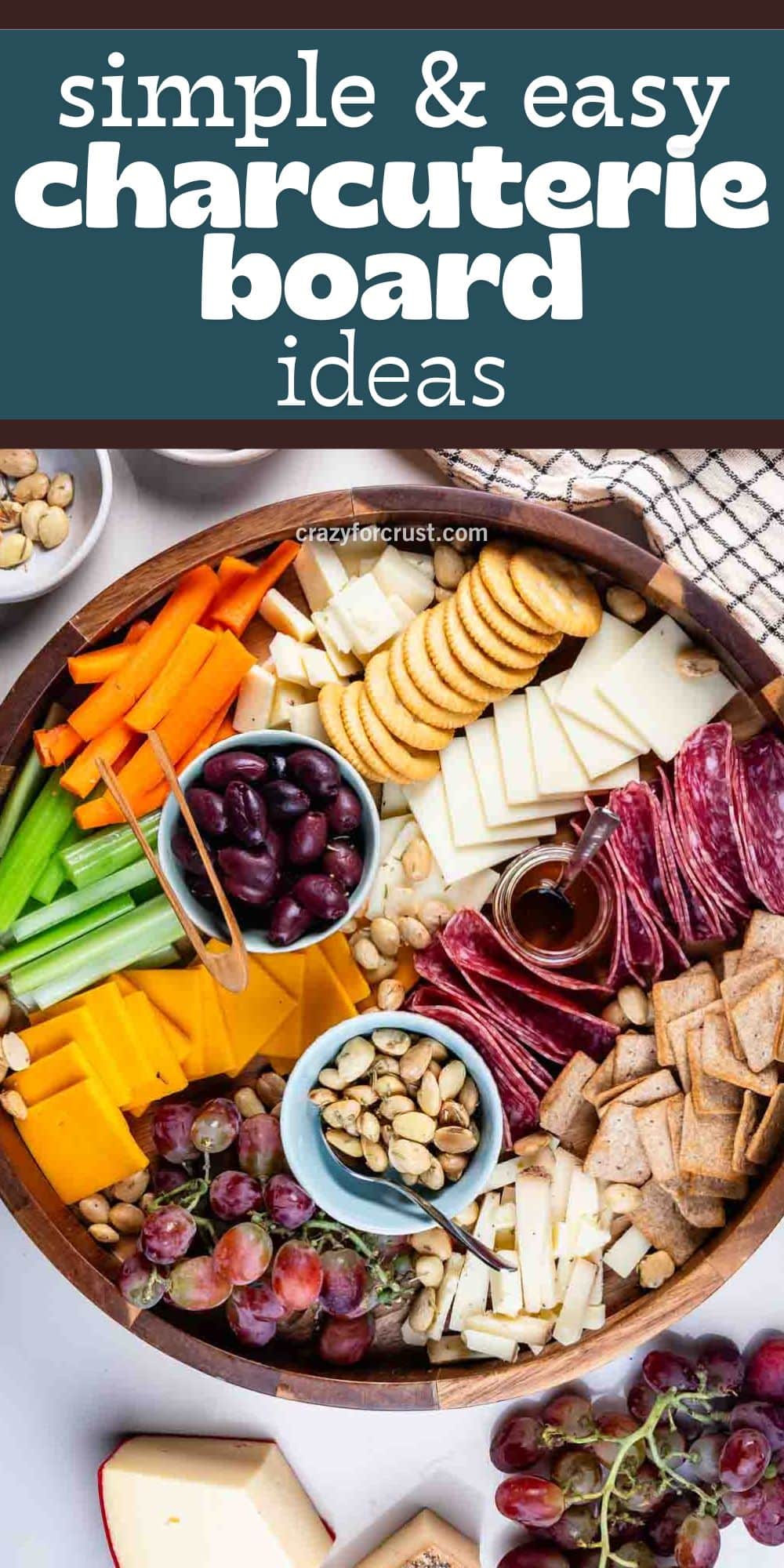 A round cheese board features sliced meats, cheeses, crackers, vegetables, nuts, olives, and grapes arranged in sections, with small bowls holding nuts and olives.
