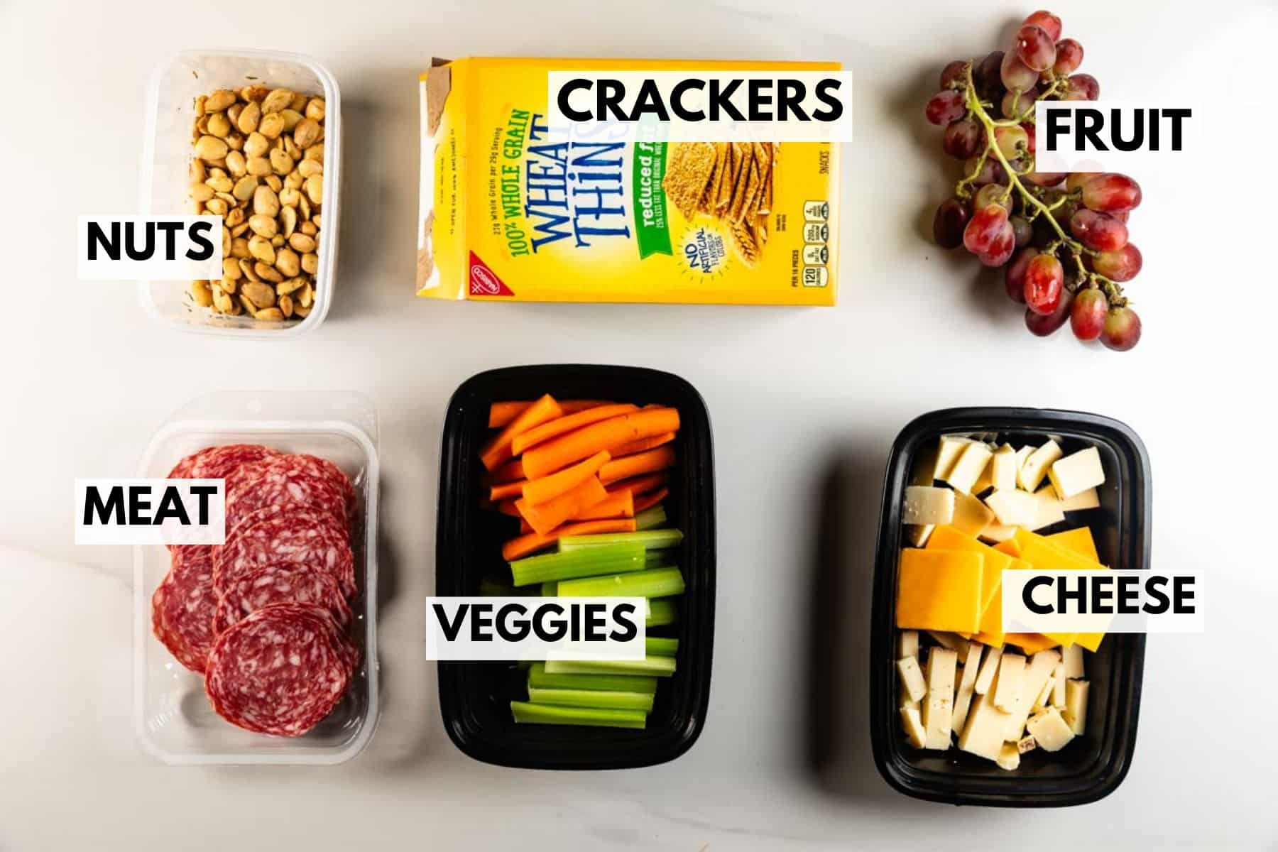 A top-down view of a cheese board shows foods in separate containers labeled: nuts, meat (salami slices), veggies (celery and carrots), crackers, cheese cubes, and a bunch of grapes labeled as fruit.