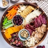 A wooden cheese board showcases assorted cheeses, crackers, sliced salami, celery, carrots, grapes, nuts, olives, and a small jar of honey. Extra bowls of nuts and olives plus a bunch of grapes surround the board for added variety.