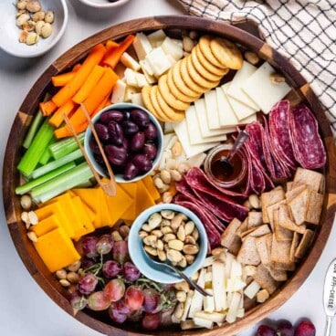 A wooden cheese board showcases assorted cheeses, crackers, sliced salami, celery, carrots, grapes, nuts, olives, and a small jar of honey. Extra bowls of nuts and olives plus a bunch of grapes surround the board for added variety.