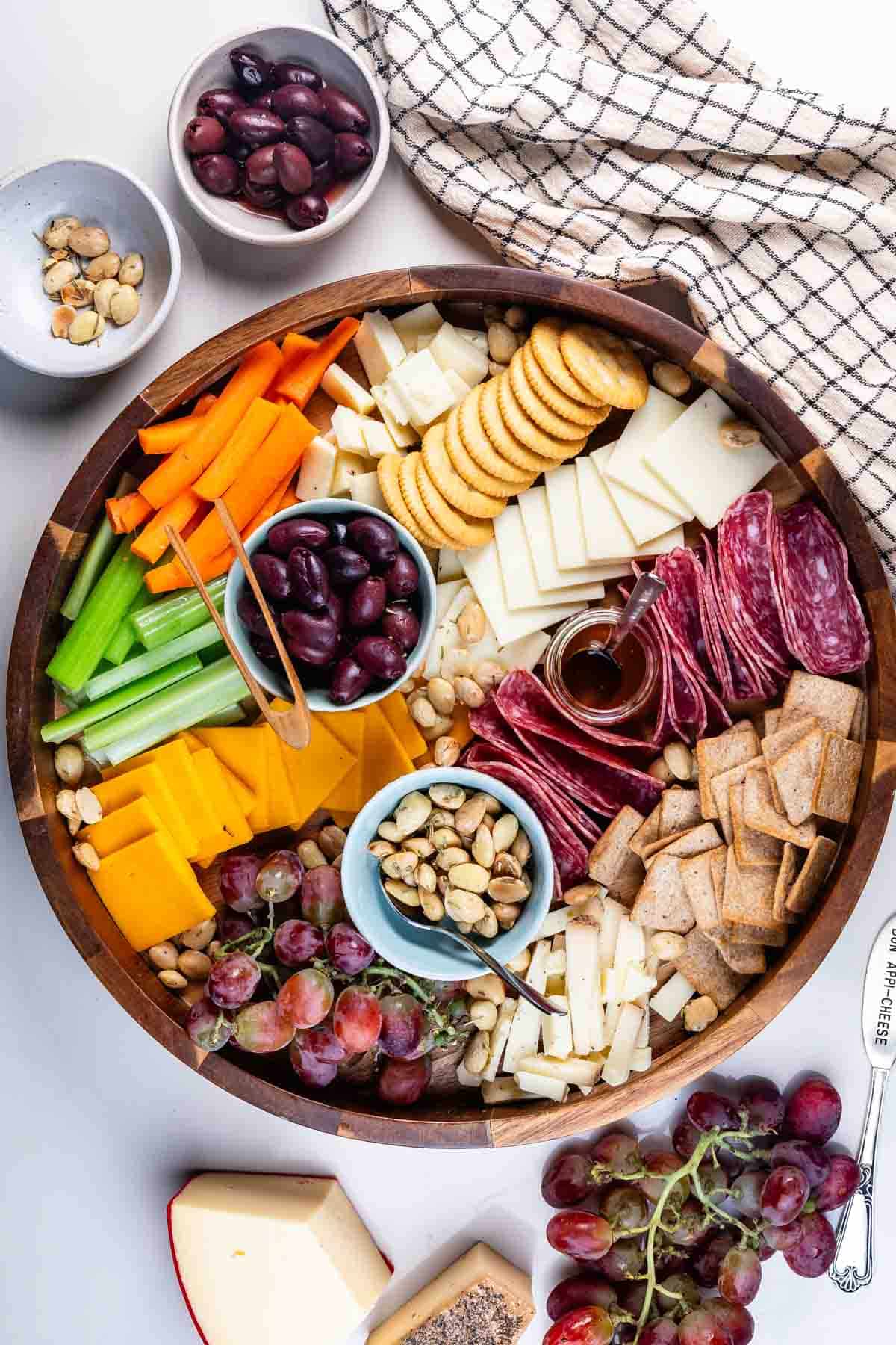 A wooden cheese board showcases assorted cheeses, crackers, sliced salami, celery, carrots, grapes, nuts, olives, and a small jar of honey. Extra bowls of nuts and olives plus a bunch of grapes surround the board for added variety.