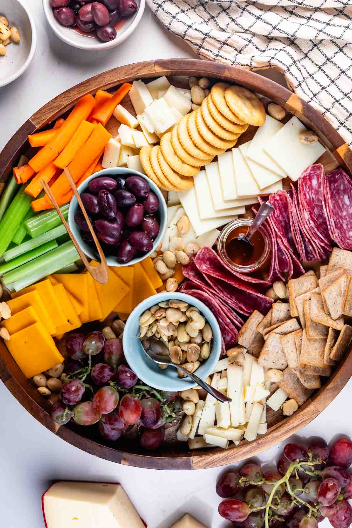 A wooden cheese board filled with assorted cheeses, crackers, sliced meats, olives, celery, carrot sticks, grapes, and a small bowl of honey. Pistachios and other nuts are also included. A checkered cloth is visible in the background.