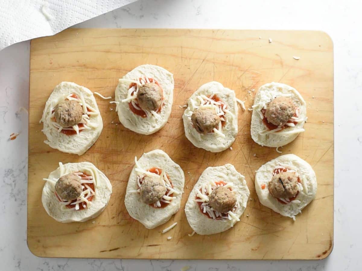 biscuits on cutting board topped with sauce, cheese, and a meatball.