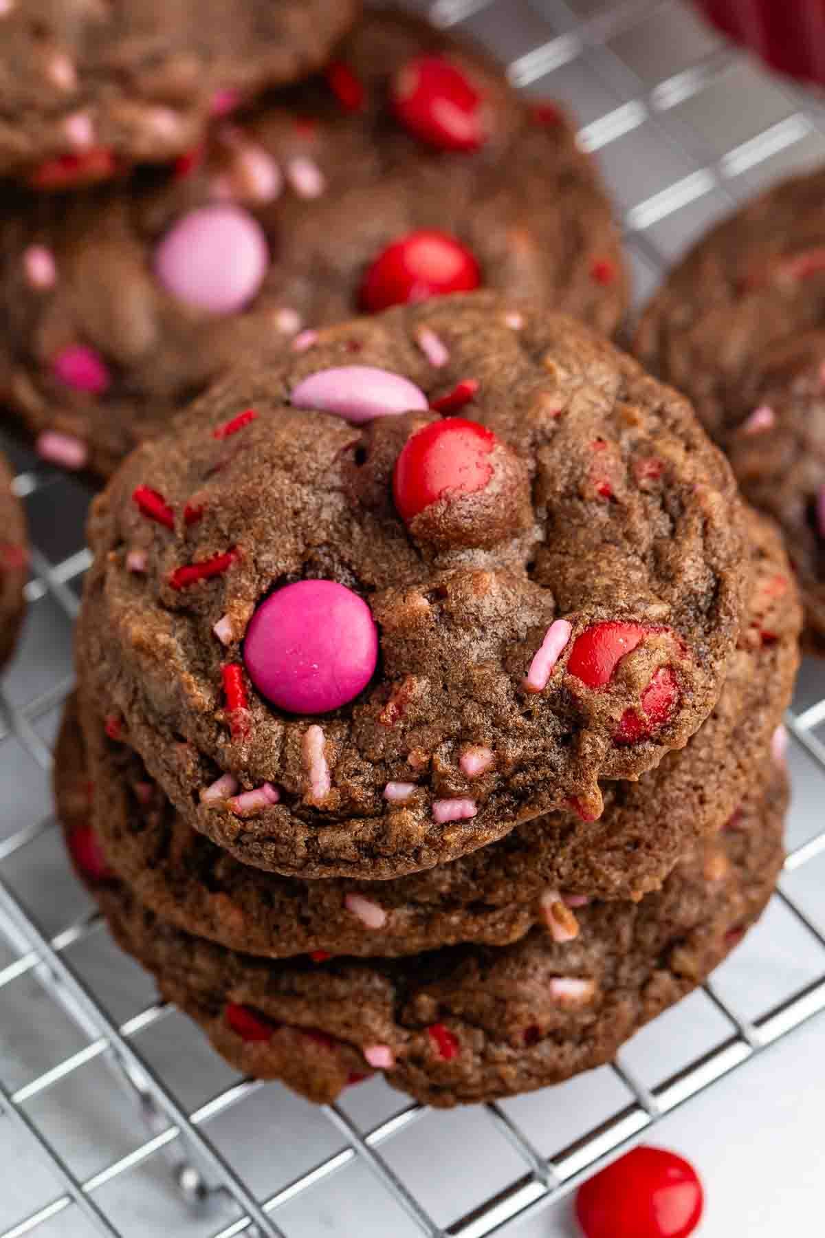 A stack of Valentine Cookies with red, pink, and purple candy-coated chocolates and sprinkles rests on a metal cooling rack.