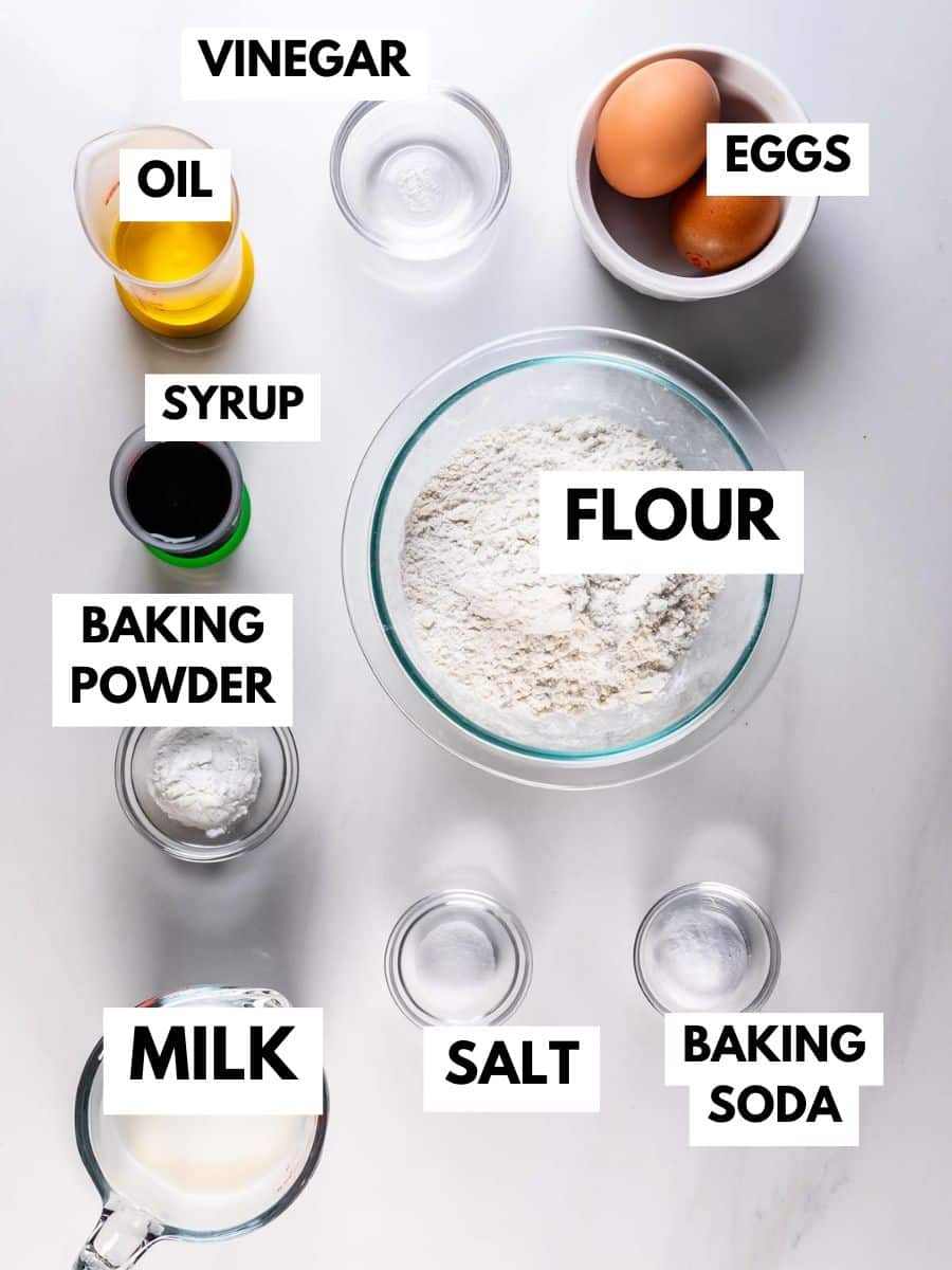 A top-down view of baking ingredients for Pancake Muffins, each labeled—eggs, flour, milk, oil, syrup, vinegar, baking powder, baking soda, and salt—arranged neatly in bowls and cups on a white surface.